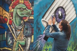 A person photographing a colorful street mural of a stylized figure with a smartphone.
