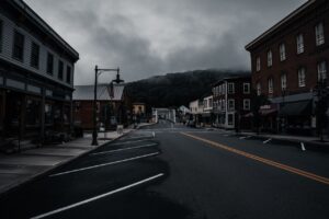 Empty small-town Main Street lined with brick buildings under a moody, fog-covered sky, with a steel truss bridge visible in the distance.