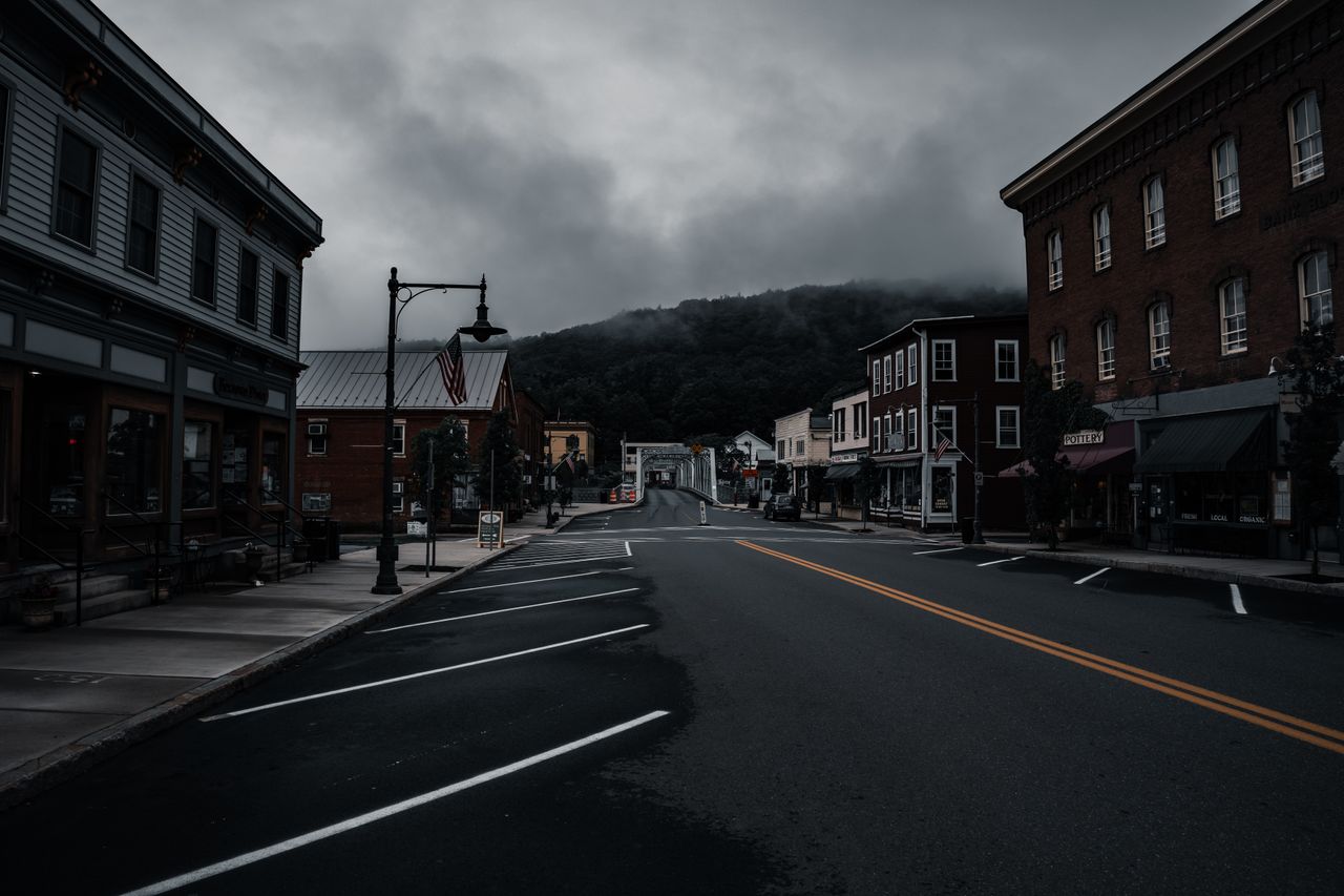 Empty small-town Main Street lined with brick buildings under a moody, fog-covered sky, with a steel truss bridge visible in the distance.